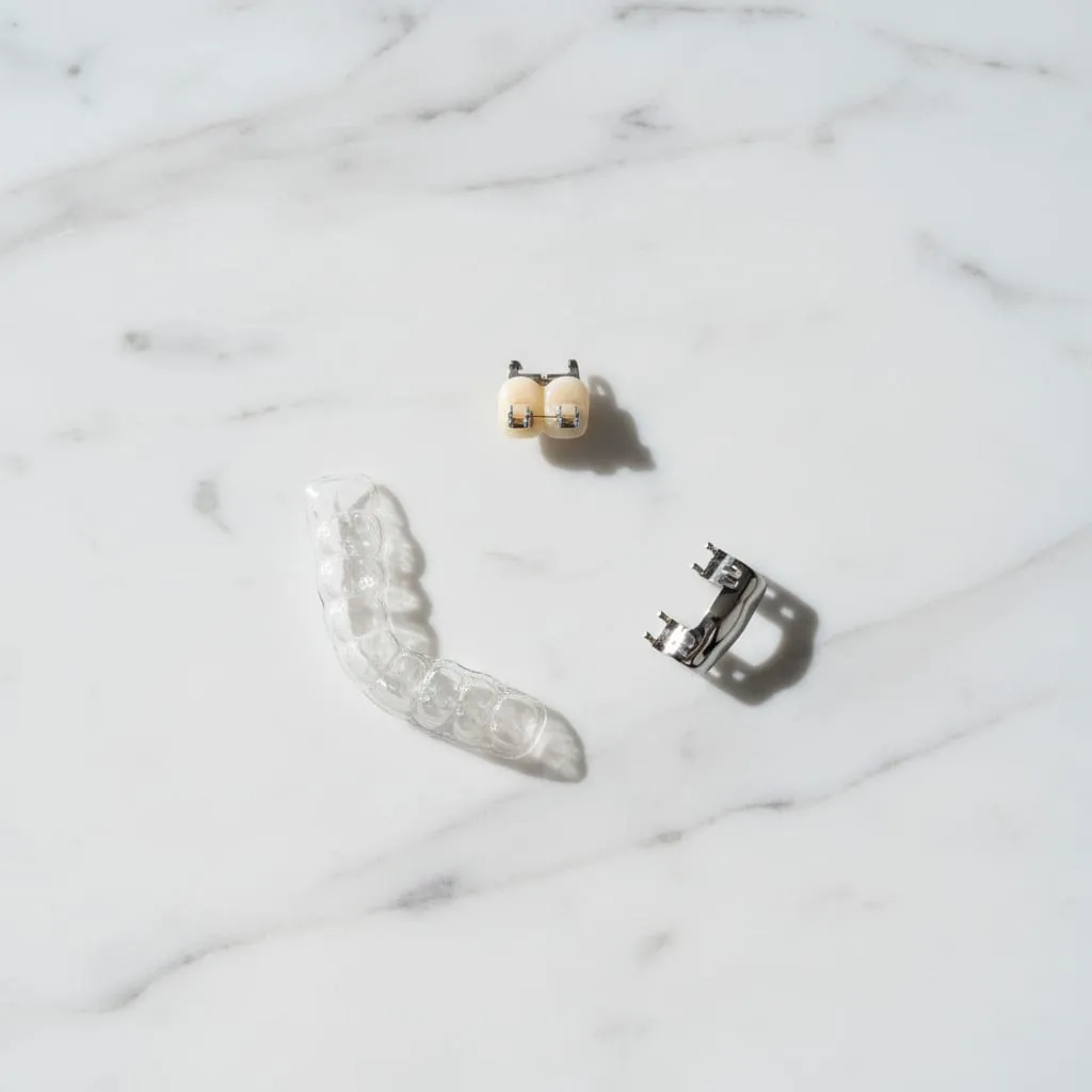 A clear aligner tray beside a ceramic bracket and a metal bracket on white marble.