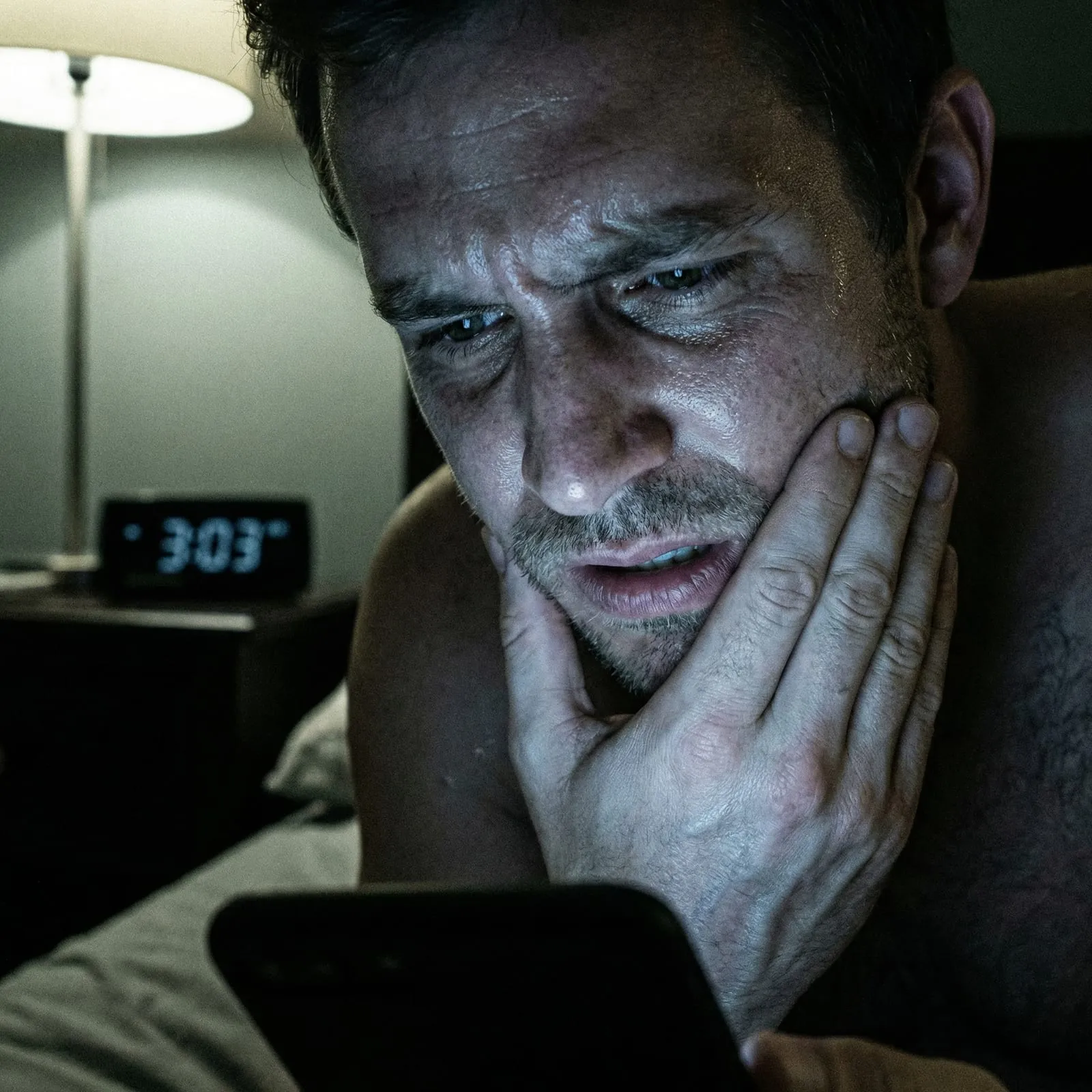 A man holds his jaw in pain at night, lit by his phone, a clock reading 3:03 behind him.