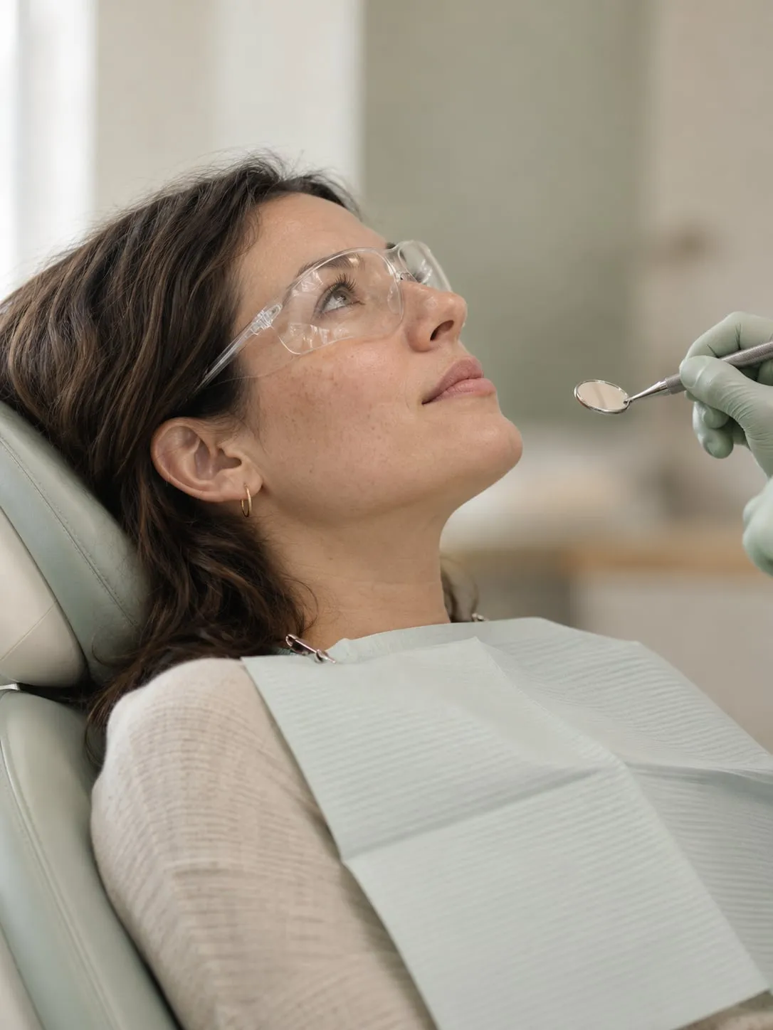 A patient in protective glasses reclines in a dental chair while a gloved hand holds a dental mirror