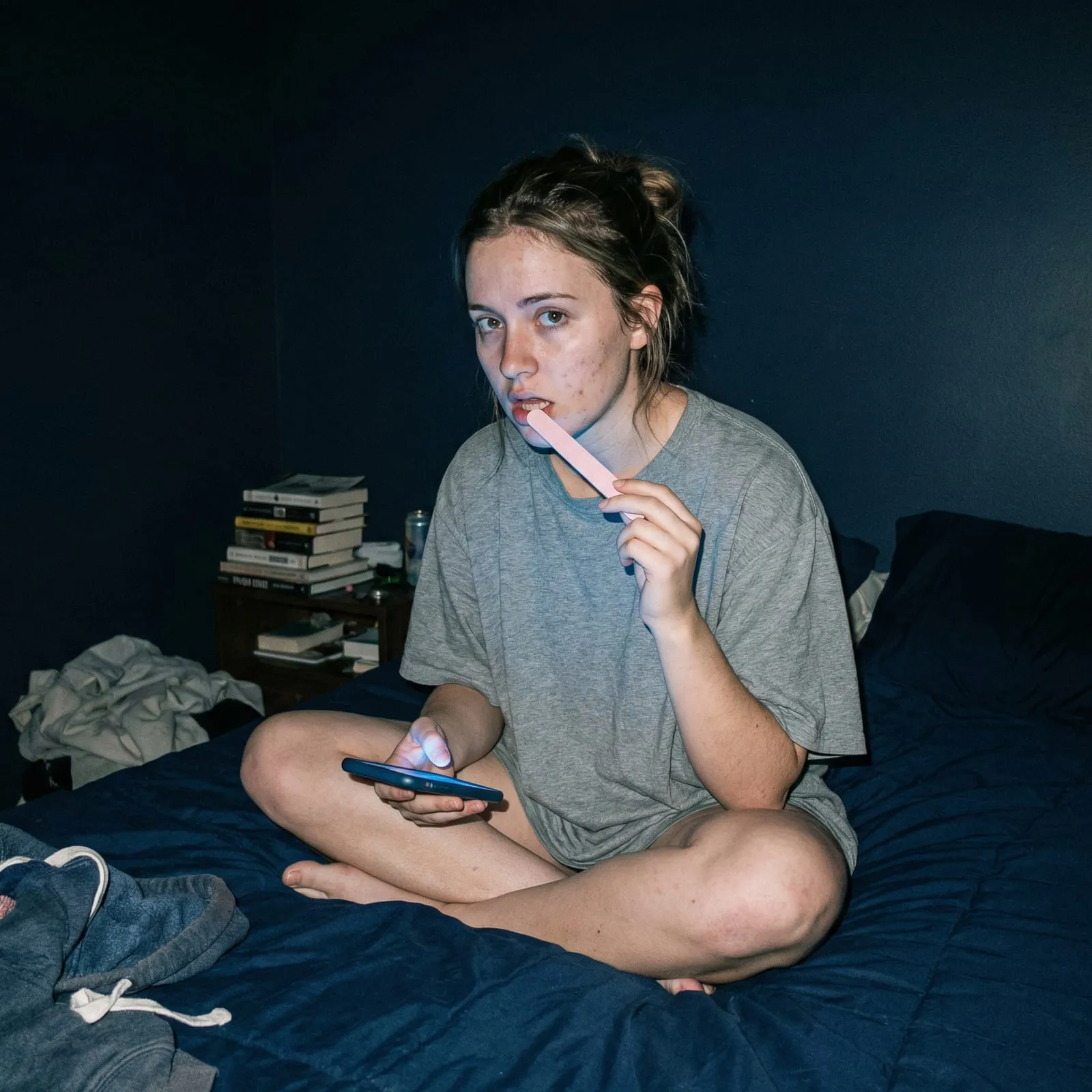 A young woman sits on her bed filing her teeth with a nail file while watching her phone.